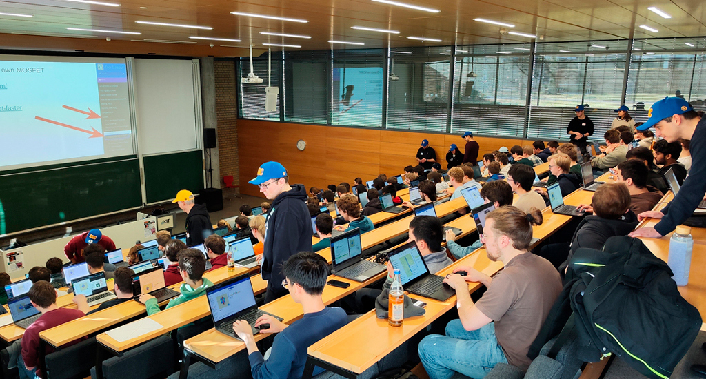 Large university lecture hall with many students using laptops, a presenter at front, and screens displaying content.